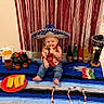 Steven is registered to the contest to win money with this photo: baby, blanket, bottle, candy, celebration, child, colorful, decorations, fiesta, food, indoors, jeans, mariachi_figurine, mexican_theme, plate, shirt, smiling, sombrero, streamers, tamales