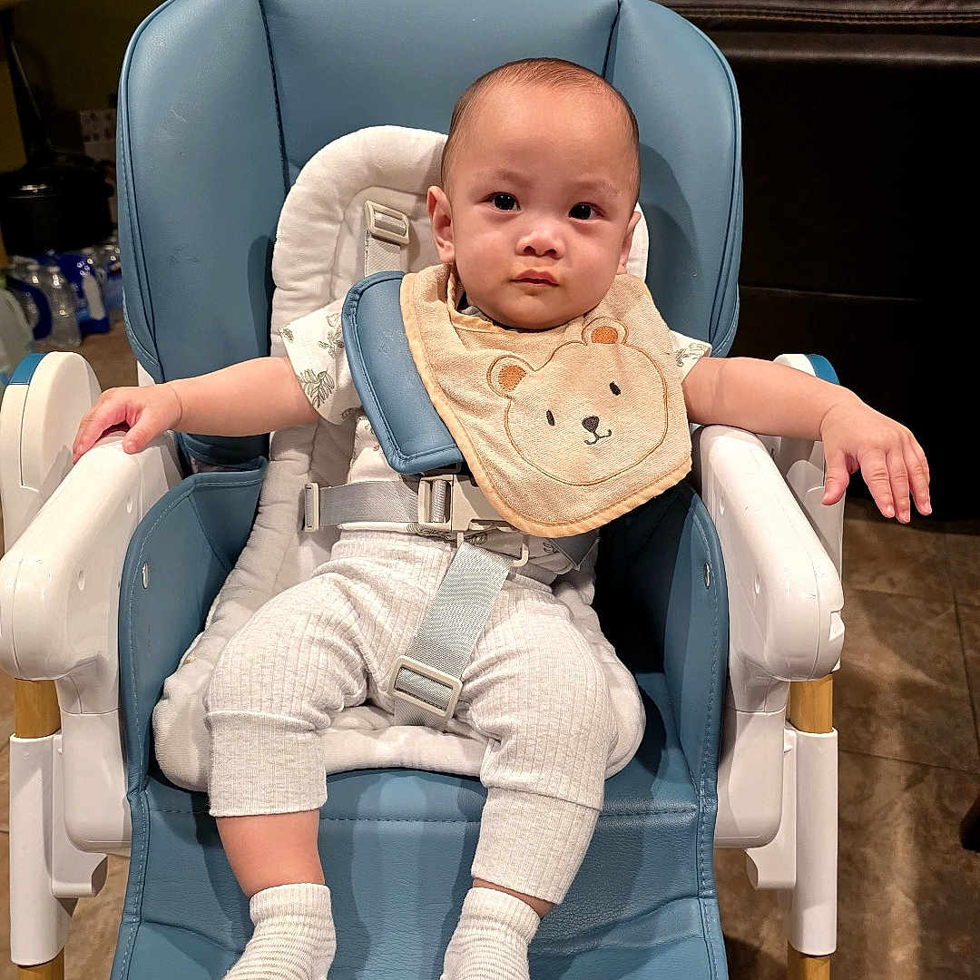 Wesley Joeward is registered to the contest to win money with this photo: baby, high_chair, bib, baby_clothes, socks, indoor, floor_tiles, furniture, child, person, seat_belt, modern_chair, home, kitchen, neutral_expression, cute, infant, white_clothing, blue_chair, toddler