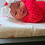 baby, infant, headband, bow, red_clothing, polka_dots, smile, face, lying_down, floral_pattern, bed, blanket, cute, indoors, person, child, adorable, soft_light, portrait, young