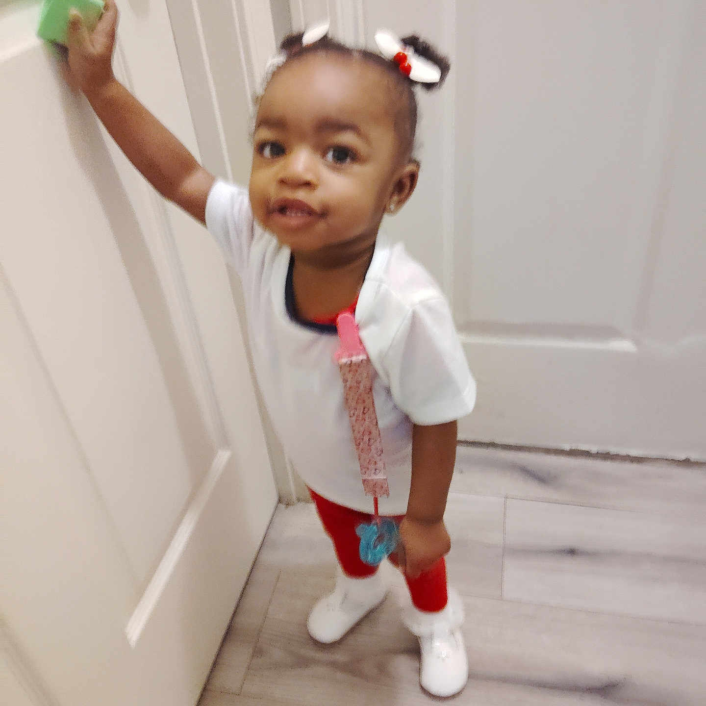 Kenyatta is registered to the contest to win money with this photo: child, curious, cute, door, door_handle, expression, floor, flooring, hair_bows, indoor, looking_up, person, playful, red_pants, small_child, standing, toddler, white_shirt, white_shoes, young_child