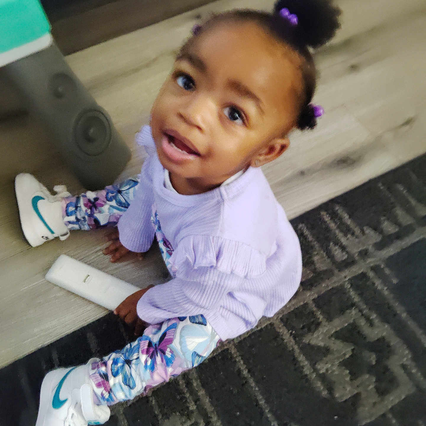 Kenyatta is registered to the contest to win money with this photo: butterfly_pattern, carpet, casual, child, curly_hair, cute, floor, hair_puffs, happy, indoor, leggings, looking_up, person, playful, purple_clothing, remote_control, sitting, toddler, white_sneakers, young_child