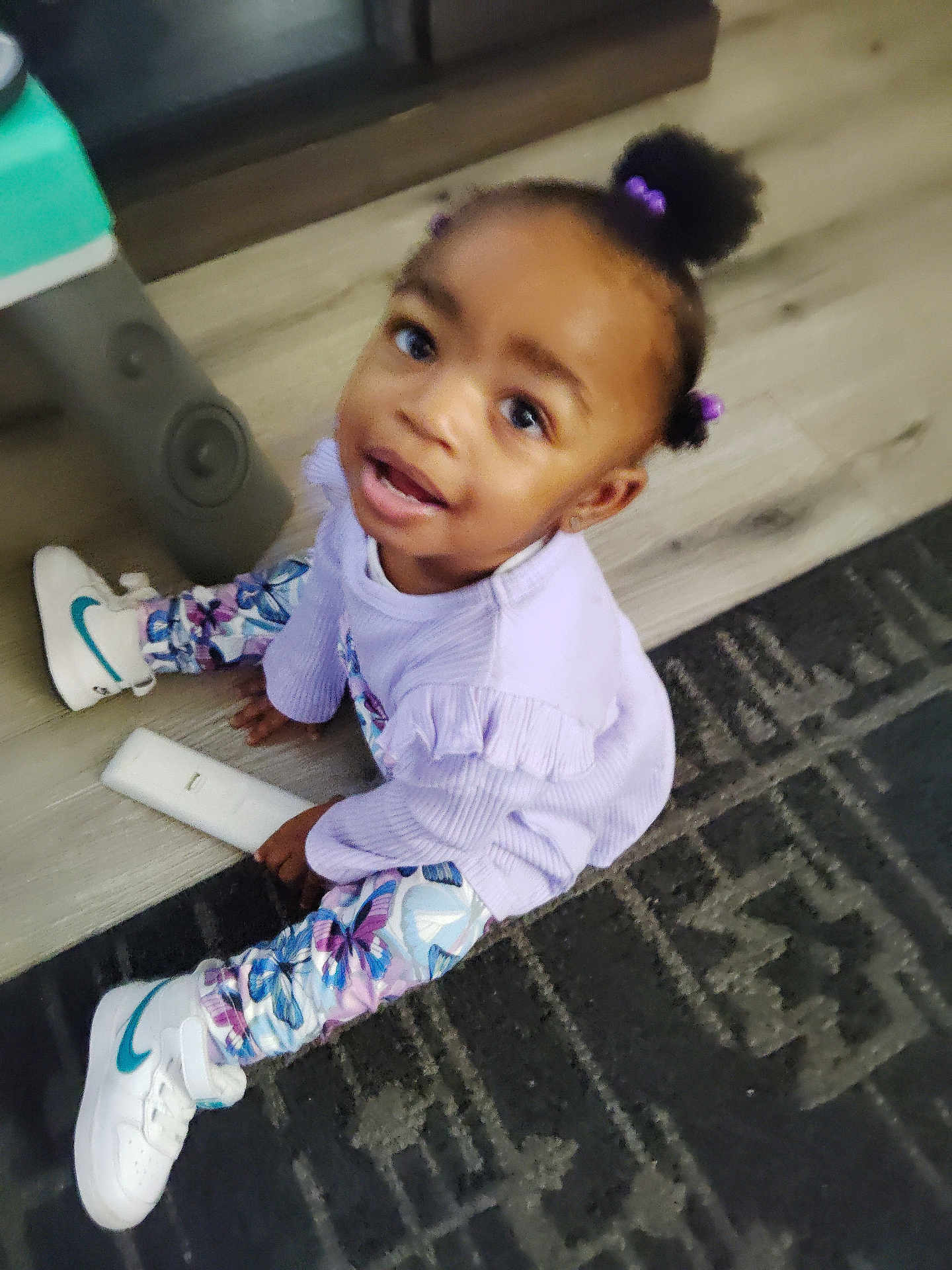 Kenyatta is registered to the contest to win money with this photo: butterfly_pattern, carpet, casual, child, curly_hair, cute, floor, hair_puffs, happy, indoor, leggings, looking_up, person, playful, purple_clothing, remote_control, sitting, toddler, white_sneakers, young_child