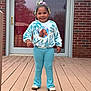 blue_pants, brick_wall, casual_clothing, child, cute, daylight, door, footwear, girl, glass_door, hair_accessories, happy, outside, person, playful, portrait, smiling, standing, sweater, wooden_deck
