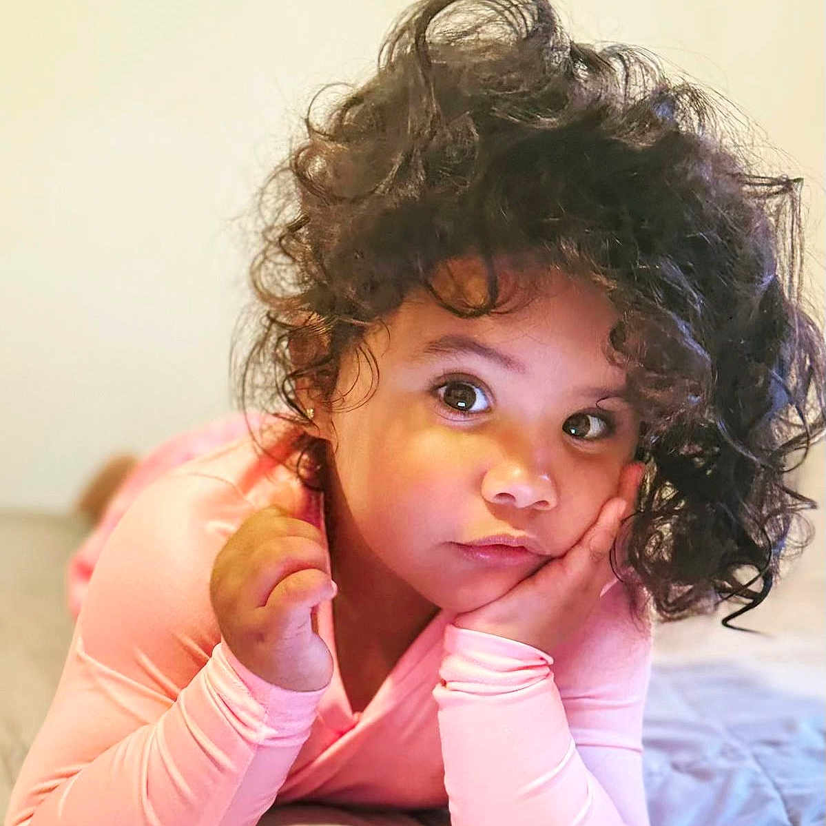 Megan Nayira joined the competition — help win amazing prizes! bed, casual, child, childhood, curious, curly_hair, cute, expression, eyes, face, hand, indoor, person, pink_clothing, portrait, relaxed, resting, skin, soft_light, young