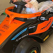 Elyna a rejoint le concours — aidez-le/la à gagner de superbes lots ! child, toddler, smile, toy_car, steering_wheel, pigtails, dress, tulle, indoor, bookshelf, toy, wheel, orange, mclaren, logos, playful, happy, portrait, floor, furniture