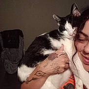 Sassy joined the competition — help win amazing prizes! cat, person, tattoo, smile, curly_hair, black_and_white_cat, indoor, shoulder, affection, closed_eyes, hand, clothing, chair, fabric, dim_light, happy, pet, feline, human, closeup