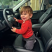 Beau is registered to the contest to win money with this photo: toddler, child, car_interior, steering_wheel, black_leather_seat, red_sweater, smile, happy, cute, baby, dashboard, window, seatbelt, car_floor_mat, indoor, portrait, young_child, car_door, natural_light, casual_clothing