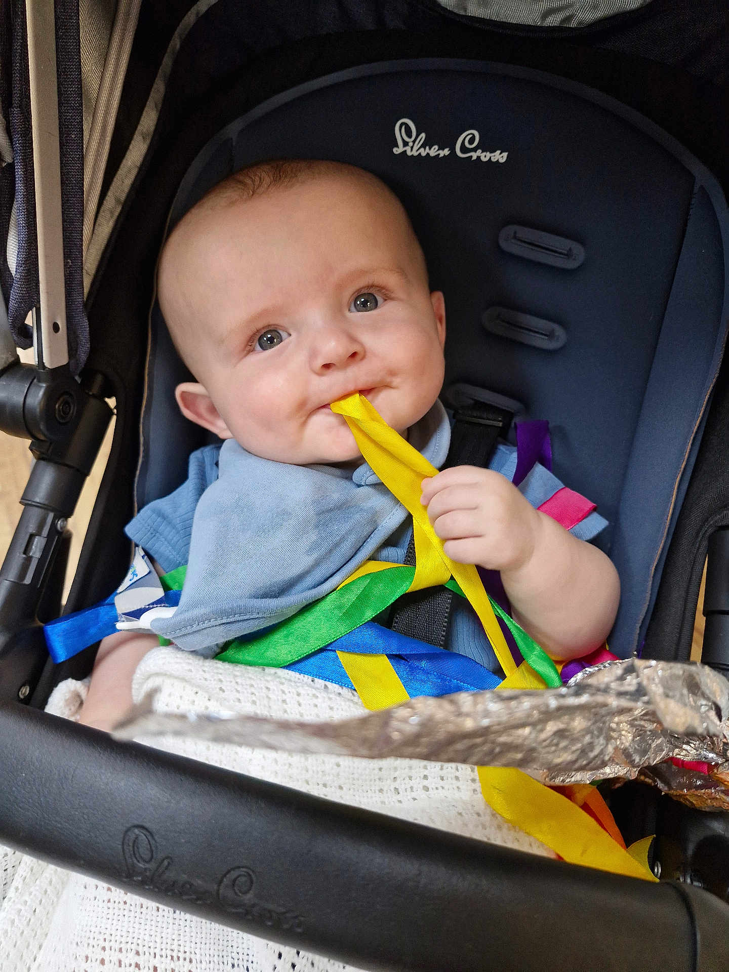 Daniel is registered to the contest to win money with this photo: baby, stroller, ribbons, yellow_ribbon, blue_bib, blanket, child, infant, seat, hand, face, chewing, colorful, toy, cute, smiling, person, clothing, indoor, soft_lighting