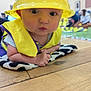 baby, child, helmet, yellow, vest, indoor, floor, wood, mat, pattern, person, curious, expression, wide_eyes, blurred_background, people, soft, clothing, infant, laying
