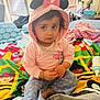 toddler, child, pink_hoodie, mouse_ears, bow, sitting, colorful_blanket, baby_toys, bottle, indoor, bed, blanket, curious_expression, soft_lighting, casual_clothing, person, face, young_child, cute, cozy
