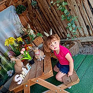 Mylana a rejoint le concours — aidez-le/la à gagner de superbes lots ! child, girl, pink_shirt, blue_shorts, wooden_bench, wooden_table, bunny_statues, flowers, potted_plants, greenery, rustic_wall, decor, playful, spring, outdoor, grass_carpet, curly_hair, earrings, garden, bench
