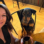 Uta participe au concours pour gagner de l'argent avec cette photo : dog, doberman, black_and_tan, pet, animal, person, woman, face, portrait, indoor, restaurant, dining_area, table, chair, wood_floor, metal_cup, drinkware, seated, selfie, warm_lighting