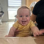 adult_hand, baby, blue_eyes, blurred_background, child, cute, expression, face, furniture, happy, indoor, infant, light, person, short_hair, skin, smiling, table, tongue_out, yellow_shirt