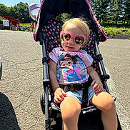 Bailee is registered to the contest to win money with this photo: toddler, sunglasses, stroller, outdoor, sunny_day, child, smiling, casual_clothing, shorts, tshirt, footwear, pavement, trees, building, reflection, seatbelt, happy, person, summer, recreation