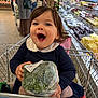 toddler, child, shopping_cart, broccoli, grocery_store, produce, happy, smiling, food, plastic_wrap, aisle, person, indoor, clothing, curly_hair, shopping, supermarket, baby, holding, retail