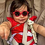 toddler, child, sunglasses, red_clothing, stroller, toy, giraffe_toy, seatbelt, baby, cute, smile, reaching_hand, infant, outdoor, sun_protection, accessory, white_shorts, playful, closeup, portrait
