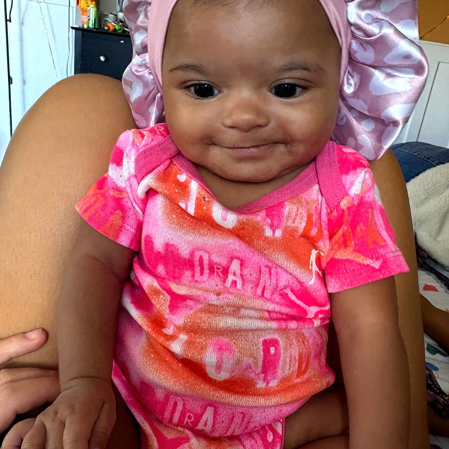 Genesis is registered to the contest to win money with this photo: baby, infant, child, pink_clothing, satin_bonnet, smile, indoors, person, cute, sitting, hand, face, skin, headwear, furniture, television, background, human, portrait, cozy