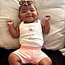 baby, infant, smiling, bow, headband, white_tank_top, pink_shorts, cushion, couch, indoor, happy, portrait, child, cute, furniture, person, skin, face, baby_clothes, relaxed