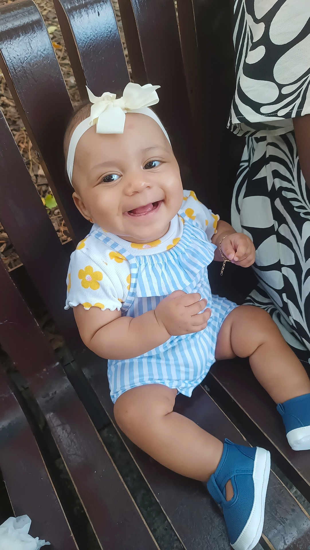 Tayla a rejoint le concours — aidez-le/la à gagner de superbes lots ! baby, child, smiling, bench, headband, bow, blue_shoes, striped_clothing, yellow_flowers, outdoor, person, cute, happy, sitting, wood, patterned_clothing, hand_bracelet, face, eyes, toes