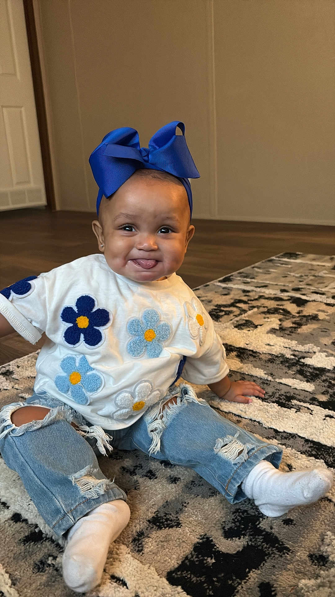 Kulture is registered to the contest to win money with this photo: baby, child, blue_bow, white_shirt, flower_patches, ripped_jeans, socks, carpet, indoor, smiling, tongue_out, cute, happy, sitting, person, floor, home, playful, young_child, casual_clothing