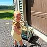 toddler, child, toy_cart, outdoor, gravel, sunlight, shadow, house, garage_door, siding, footwear, shorts, shirt, play, daylight, person, summer, greenery, wooden_toy, curious