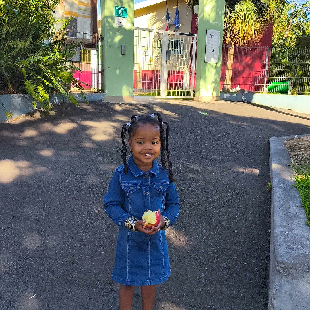Jayani participe au concours pour gagner de l'argent avec cette photo : apple, casual, child, daytime, denim_dress, gate, girl, greenery, happy, outdoor, pavement, pigtails, plants, portrait, sandals, school, shadow, smiling, sunlight, trees