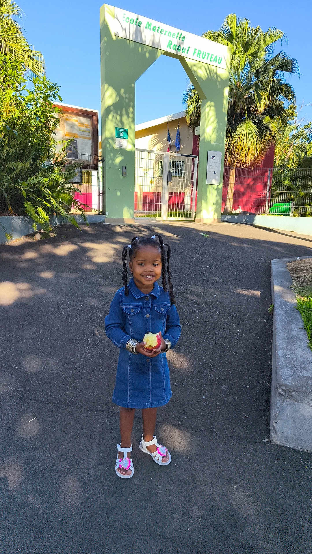 Jayani participe au concours pour gagner de l'argent avec cette photo : child, girl, denim_dress, apple, smiling, pigtails, sandals, outdoor, school, gate, pavement, trees, sunlight, shadow, plants, greenery, happy, daytime, casual, portrait
