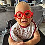 Stanislas participe au concours pour gagner de l'argent avec cette photo : baby, child, glasses, red_glasses, high_chair, bib, hand_holding, living_room, sofa, television, smiling, blond_hair, cute, indoor, person, infant, furniture, happy, face, closeup