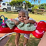 Kaniela-Ray is registered to the contest to win money with this photo: baby, child, outdoor, sunny, grass, red_baby_jumper, blanket, smiling, happy, toys, feet, face, playtime, person, greenery, palm_trees, car, chair, daytime, park