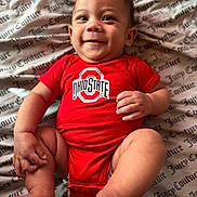 Kaniela-Ray joined the competition — help win amazing prizes! baby, smiling, red_clothing, onesie, infant, cute, lying_down, hand, foot, blanket, pattern, text_on_clothing, child, happy, portrait, young, skin, person, indoors, closeup