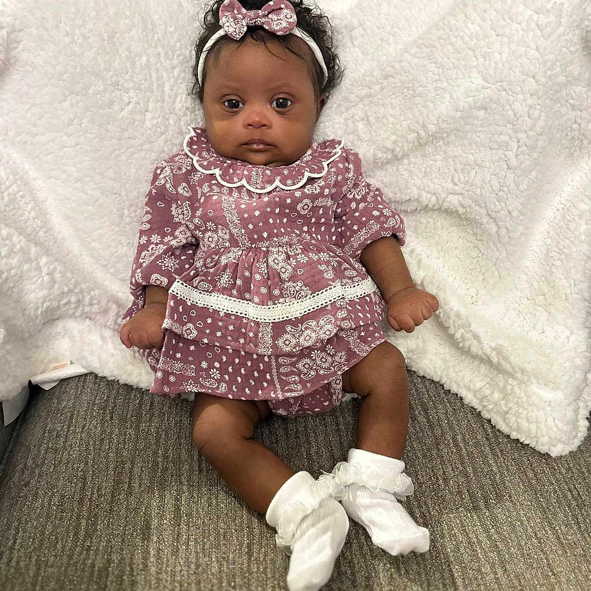 Anjali joined the competition — help win amazing prizes! baby, child, infant, portrait, sitting, indoors, blanket, carpet, headband, bow, dress, patterned_clothing, socks, white_socks, dark_skin, curly_hair, face, wide_eyes, cozy, fashion