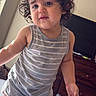Valentina is registered to the contest to win money with this photo: toddler, child, curly_hair, striped_romper, standing, portrait, indoors, home, sunlight, window_blinds, television, dresser, wood_furniture, carpet, adult_present, close_up, facial_expression, brown_hair, casual_clothing, family_scene