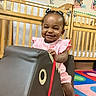 child, toddler, girl, smiling, portrait, classroom, daycare, crib, chair, furniture, pink_outfit, hair_bows, socks, colorful_rug, indoor, playroom, happy, sitting, standing, phone_screenshot
