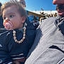 baby, pacifier, beaded_chain, jacket, child, adult, outdoor, blue_sky, person, face, hand, clothing, holding, portrait, daylight, casual, cute, warm_clothing, people, background_blur
