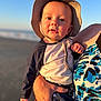 baby, child, hat, beach, sunlight, adult_hand, clothing, stars, outdoor, portrait, summer, ocean, sky, holding, casual_wear, sun_hat, face, person, warm_light, expression