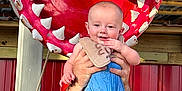 Braxton is registered to the contest to win money with this photo: adult_hands, baby, blue_clothing, child, cute, happy, holding, human, infant, jaws, outdoor, playful, portrait, red_background, rope, shark_mouth, smiling, sunlight, toy, wooden_structure