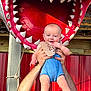 baby, child, adult_hands, shark_mouth, jaws, red_background, blue_clothing, smiling, outdoor, sunlight, rope, wooden_structure, playful, cute, portrait, holding, happy, infant, human, toy