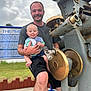 man, baby, smiling, tongue_out, outdoor, metal_machine, rust, grass, sign, cloudy_sky, shorts, tshirt, holding, happy, person, father, child, playful, nature, daytime