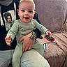 baby, child, man, tattoo, hoodie, smile, couch, living_room, onesie, hand, person, indoor, casual_clothing, happy, seated, technology, remote_control, charger, cord, portrait