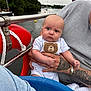 baby, infant, person, tattoo, hand, boat, water, lake, sailboat, life_buoy, clothing, expression, outdoor, adult, bracelet, nature, sky, trees, face, holding