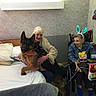 Beaowulf is registered to the contest to win money with this photo: elderly_woman, german_shepherd, dog, hospital_bed, wheelchair, bunny_ears, snacks, gifts, indoor, smiling, gray_hair, blanket, pillow, table, chair, medical_room, joy, companion, friendship, care