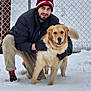 man, dog, golden_retriever, snow, winter_clothing, beanie, gloves, chain_link_fence, outdoor, pet, happy, smiling, crouching, cold_weather, pants, sneakers, fur, tail, canine, friendship