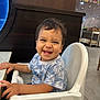 toddler, child, smiling, high_chair, blue_shirt, leaf_pattern, indoor, restaurant, table, happy, person, face, hair, seat, furniture, cute, baby, expression, joy, hands