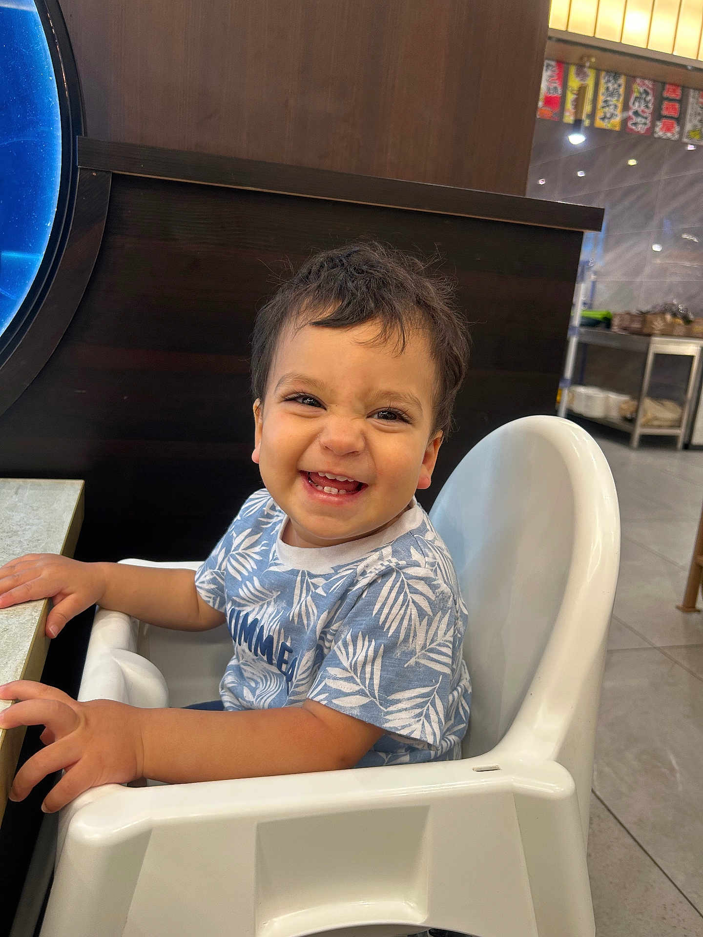 Ali participe au concours pour gagner de l'argent avec cette photo : toddler, child, smiling, high_chair, blue_shirt, leaf_pattern, indoor, restaurant, table, happy, person, face, hair, seat, furniture, cute, baby, expression, joy, hands