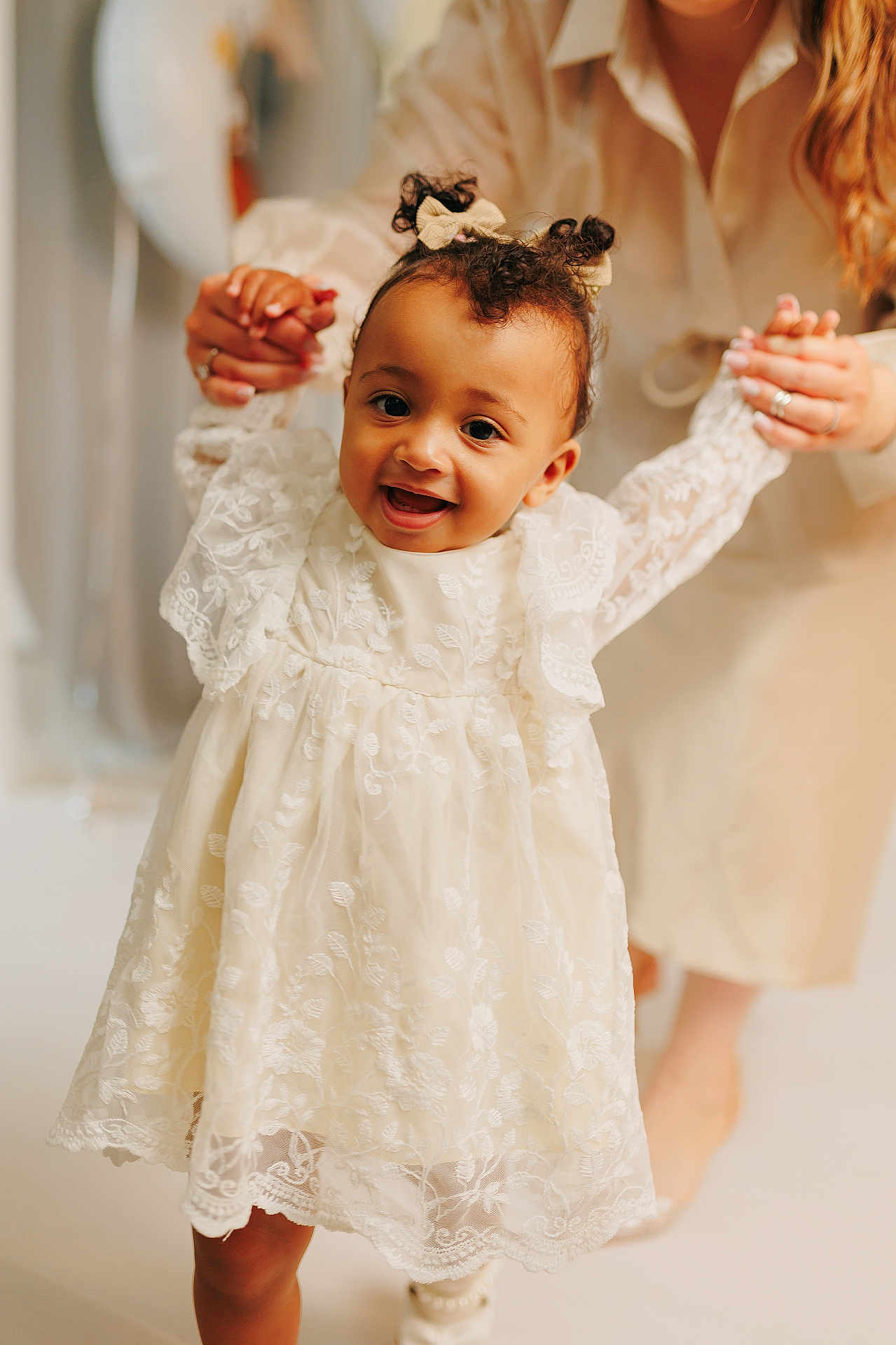 Aivi is registered to the contest to win money with this photo: adult, baby, bows, child, closeup, curly_hair, cute, footwear, hands, happy, indoor, lace, light_skin, people, portrait, smiling, supporting, toddler, walking, white_dress