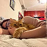 person, cat, glasses, bed, blanket, indoor, furniture, painting, cabinet, orange_cat, hugging, cozy, resting, warm_lighting, sleepy, comfort, relaxation, soft_texture, casual, intimate_moment