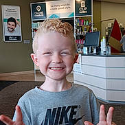 Austin is registered to the contest to win money with this photo: child, boy, smiling, peace_sign, blonde_hair, tshirt, nike, indoor, salon, hair_products, modern, counter, carpet, ceiling_lights, person, portrait, casual_clothing, happy, young, cute
