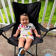 Albin is registered to the contest to win money with this photo: baby, child, smiling, sitting, chair, outdoor, porch, fence, grass, footwear, clothing, mickey_mouse, black_chair, happy, person, young_child, daylight, concrete, casual_wear, playful