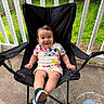 baby, child, smiling, sitting, chair, outdoor, porch, fence, grass, footwear, clothing, mickey_mouse, black_chair, happy, person, young_child, daylight, concrete, casual_wear, playful