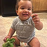 baby, child, toddler, smiling, sitting, indoor, kitchen, floor, leafy_greens, food, diaper, clothing, cabinet, appliance, curly_hair, cute, happy, person, young_child, casual